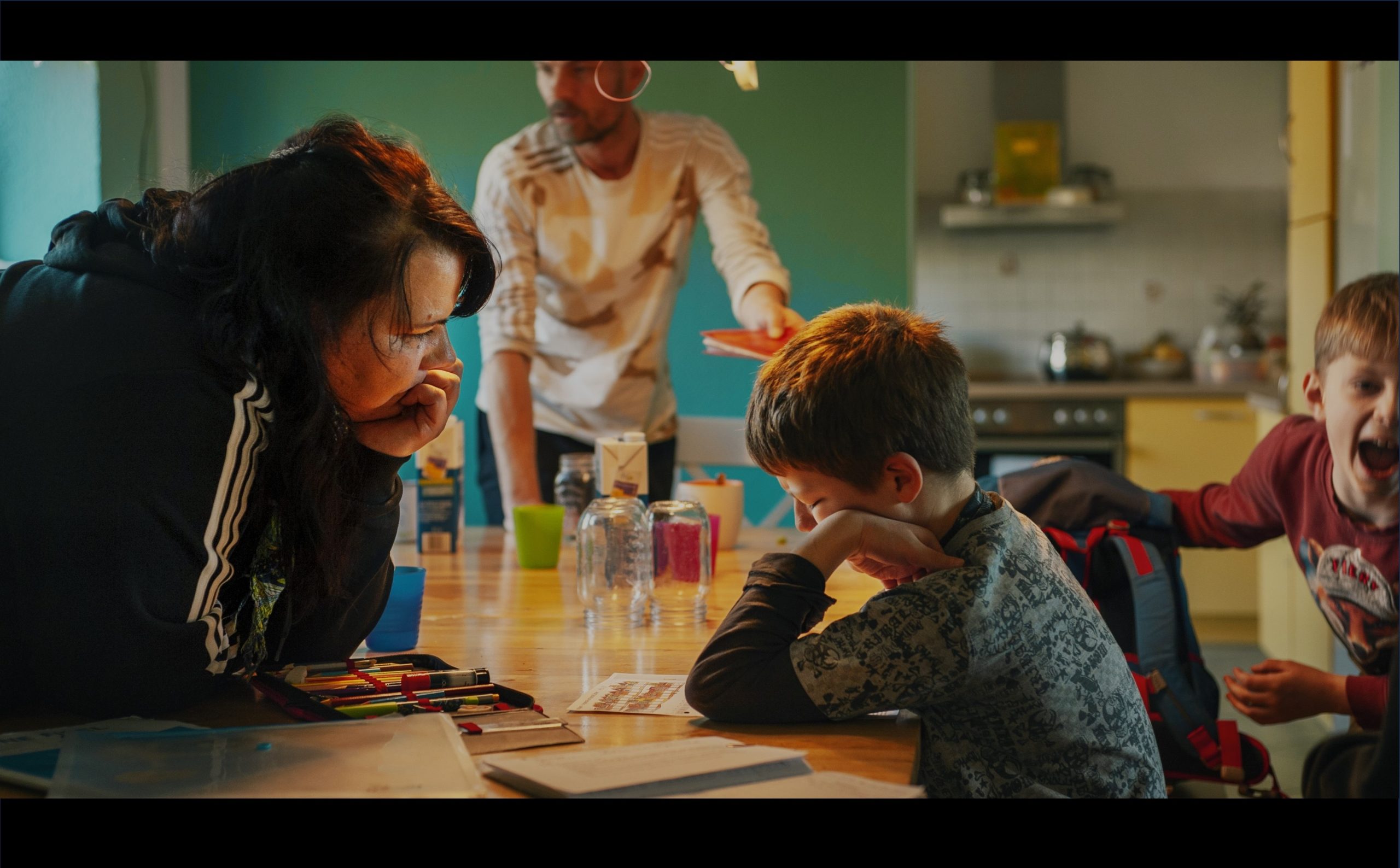 Videostill zeigt eine Frau und ein Kind am Tisch sitzen, im Hintergrund steht ein Mann