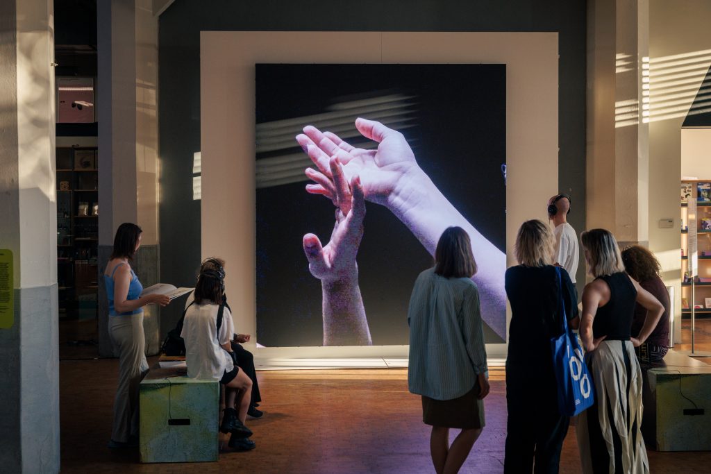 Videoinstallation im Foyer des ZKM, basierend auf der Live-Performance vom 1.8.2026. Im gezeigten Ausschnitt sind zwei sich berührende Hände zu sehen. - Copyright: Soft Signals, Davit Khorbaladze, Natia Chikvaidze, Karolina Sobel, 2026. © Stadt Karlsruhe, Kulturamt, Foto: Felix Grünschloß