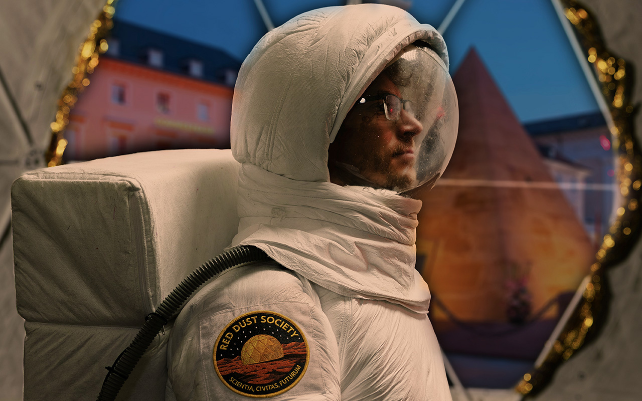 Potrait im Profil einer männlich gelesenen weißen Person mit Brille in einem weißen Astronautenanzug vor einem Gebäude